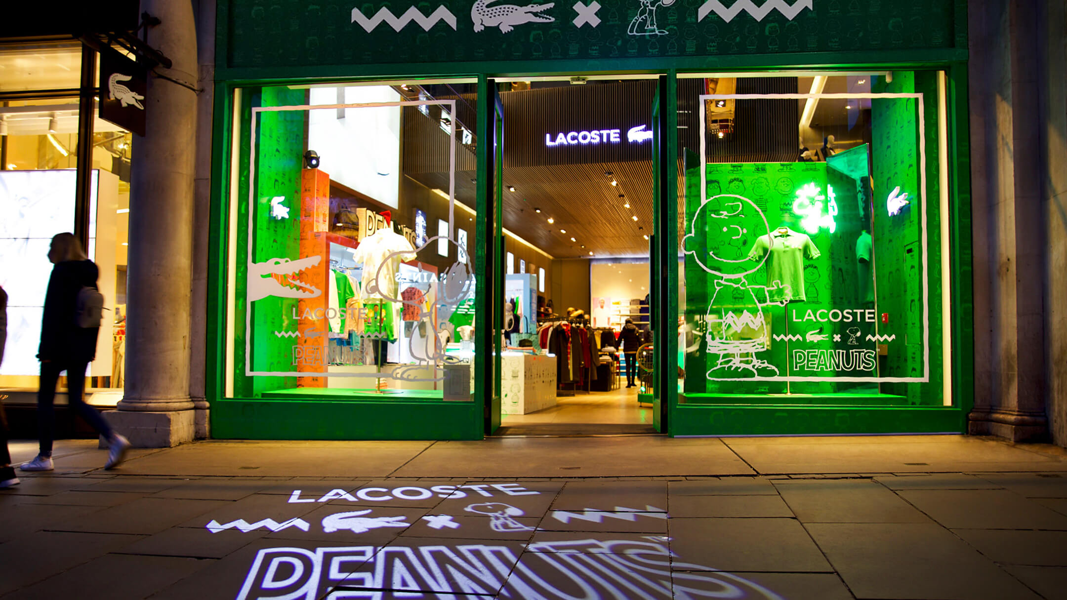 Lacoste store at night featuring Peanuts collaboration display with projected logo and illuminated interior.