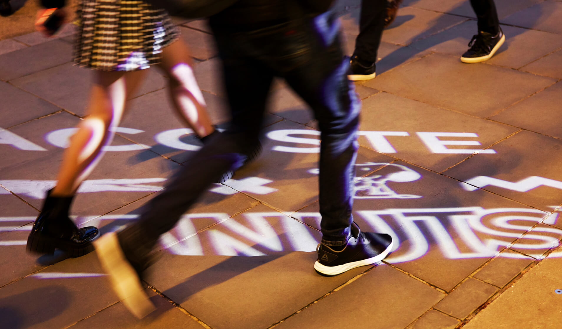 People walking across a sidewalk with a Lacoste x Peanuts logo projected in white light on the pavement.