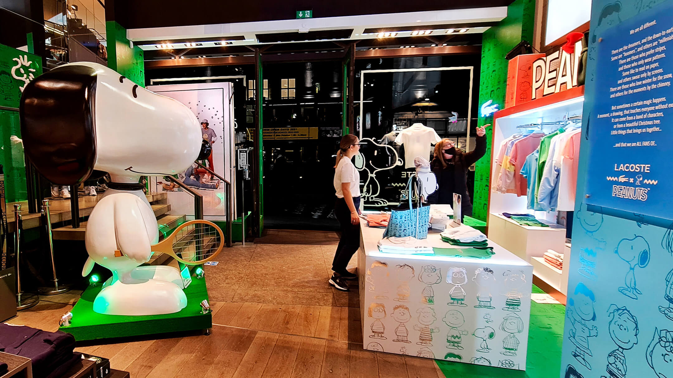 Interior of a Lacoste store featuring a large Snoopy statue, Peanuts-themed décor, and Lacoste x Peanuts merchandise on display, with staff and customers interacting.