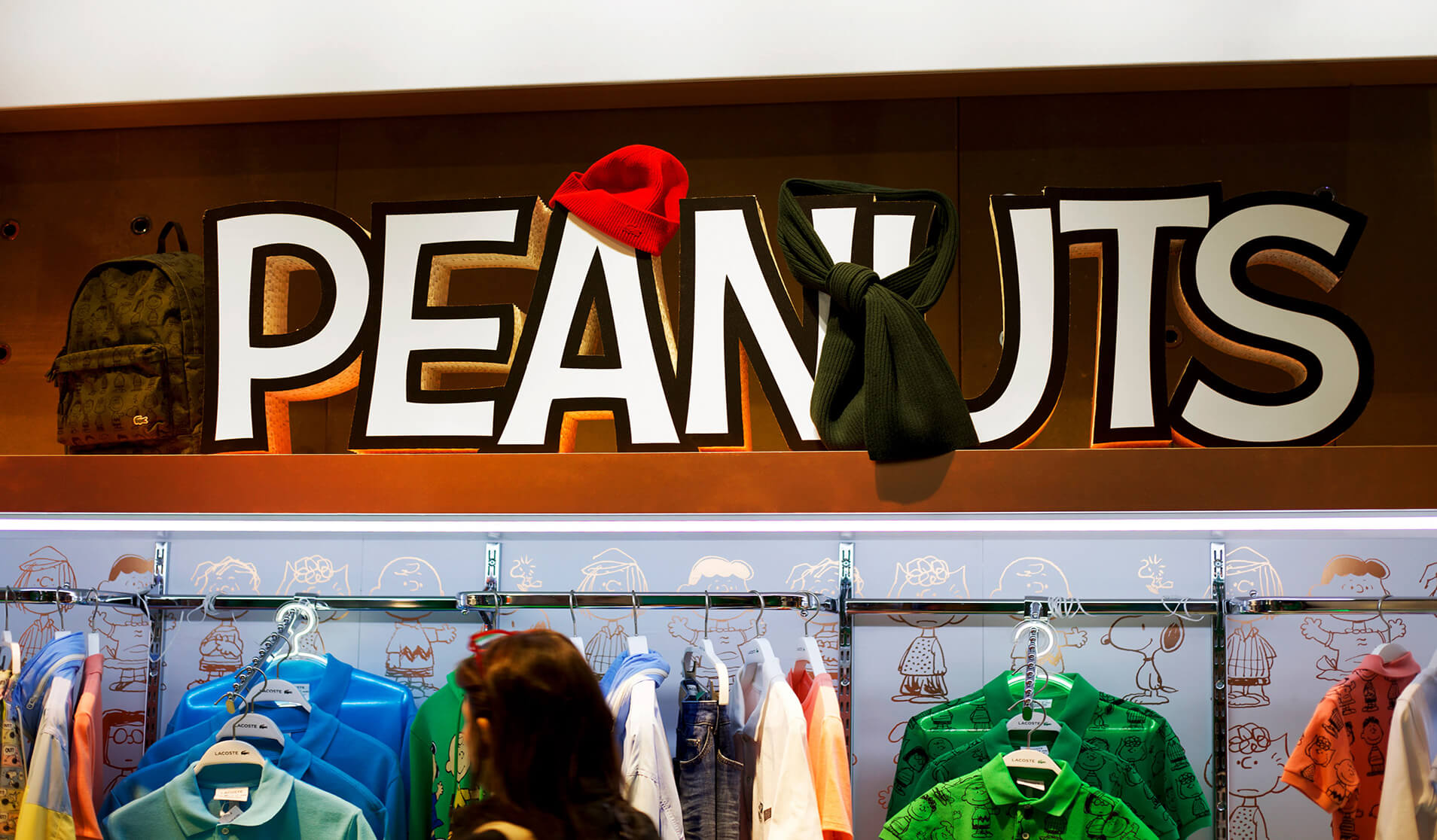 Close-up of a Peanuts sign in a Lacoste store with character hats and themed clothing displayed below.