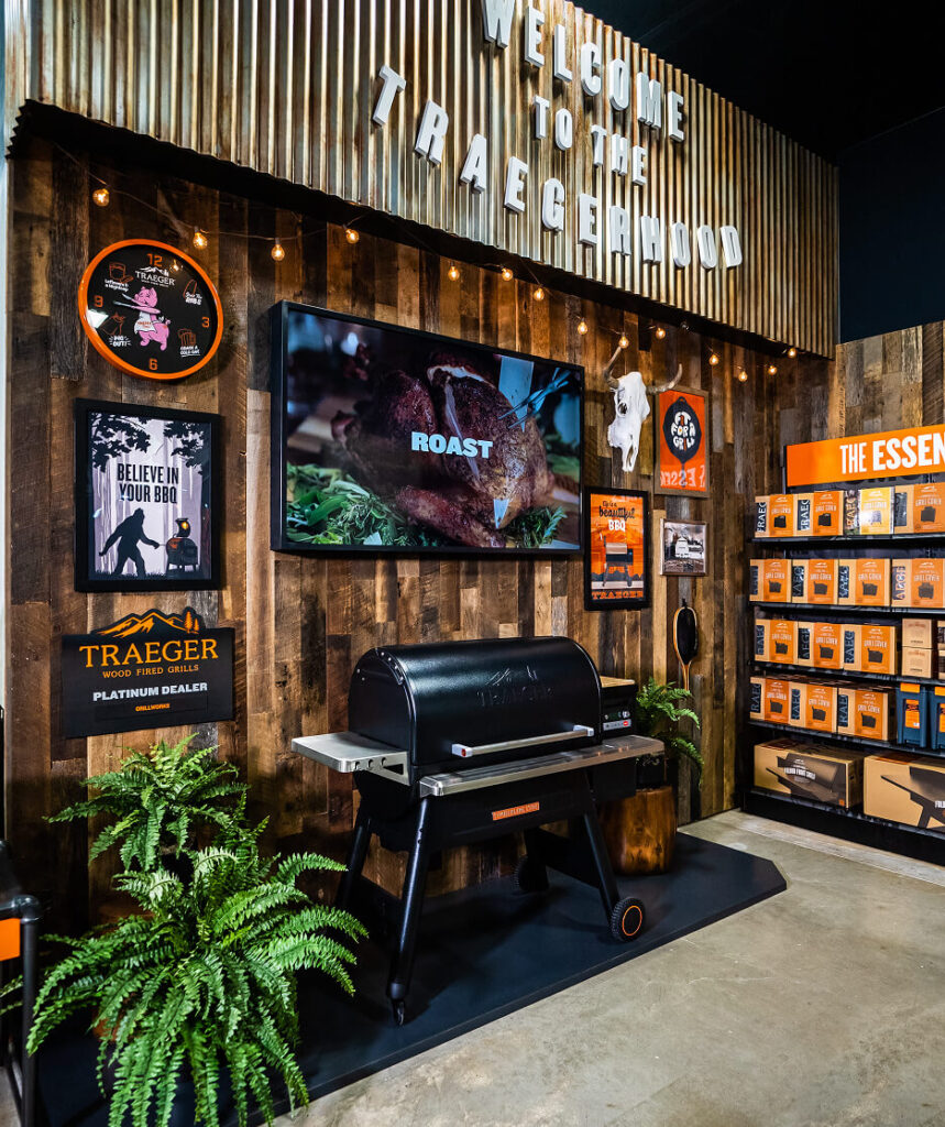 Rustic in-store Traeger display with Timberline grill, digital roast visual, wood-paneled wall, and grilling accessories.