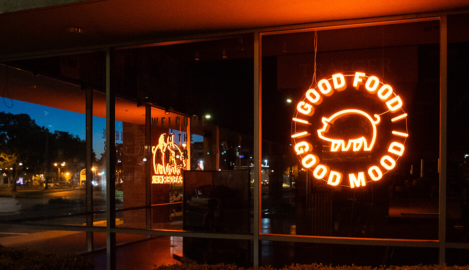 Neon pig-shaped sign in a storefront window reading “Good Food Good Mood” at night.