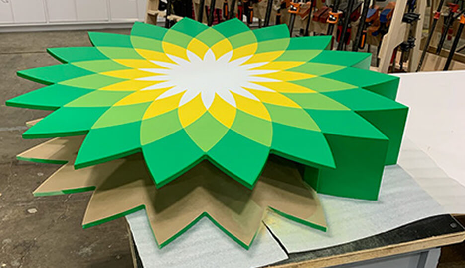 Three-dimensional BP logo sculpture in vibrant green, yellow, and white colors, layered for visual depth, placed on a workshop surface.