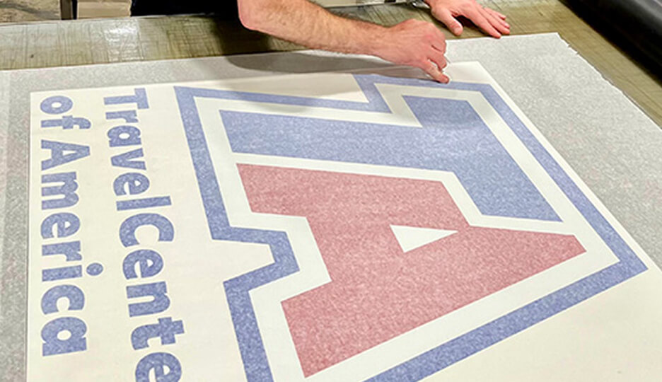 Person applying a large vinyl graphic of the TravelCenters of America (TA) logo on a flat surface, using their hand to smooth it out.