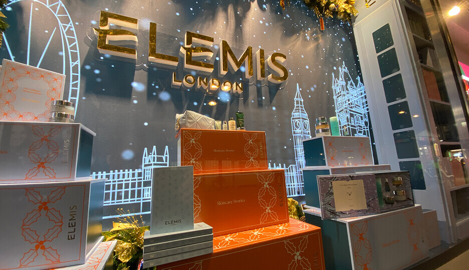 Holiday-themed Elemis London window display with stacked “Skincare Stories” gift boxes, skincare products, and illuminated London landmarks.