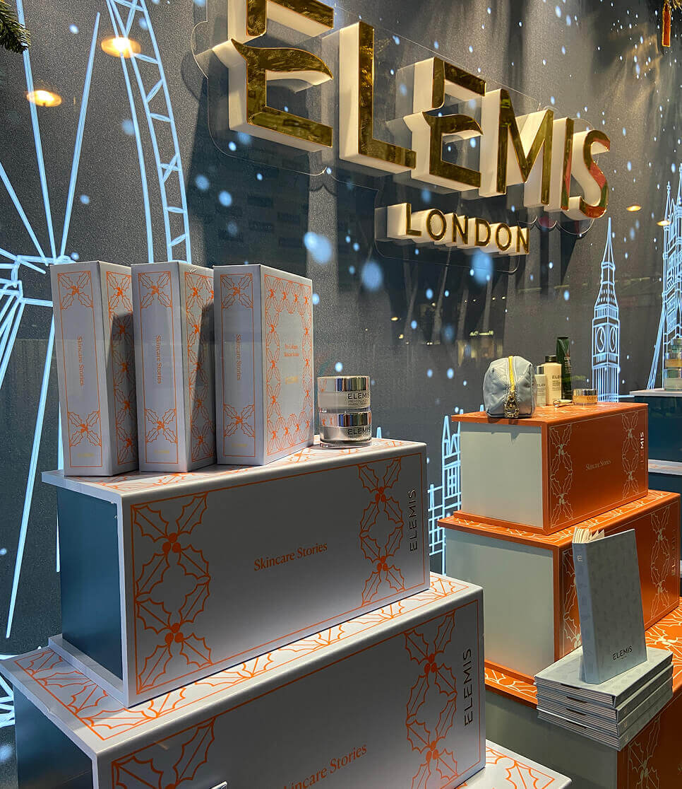 Holiday display of Elemis “Skincare Stories” gift boxes with skincare products, under a golden 3D “Elemis London” sign.