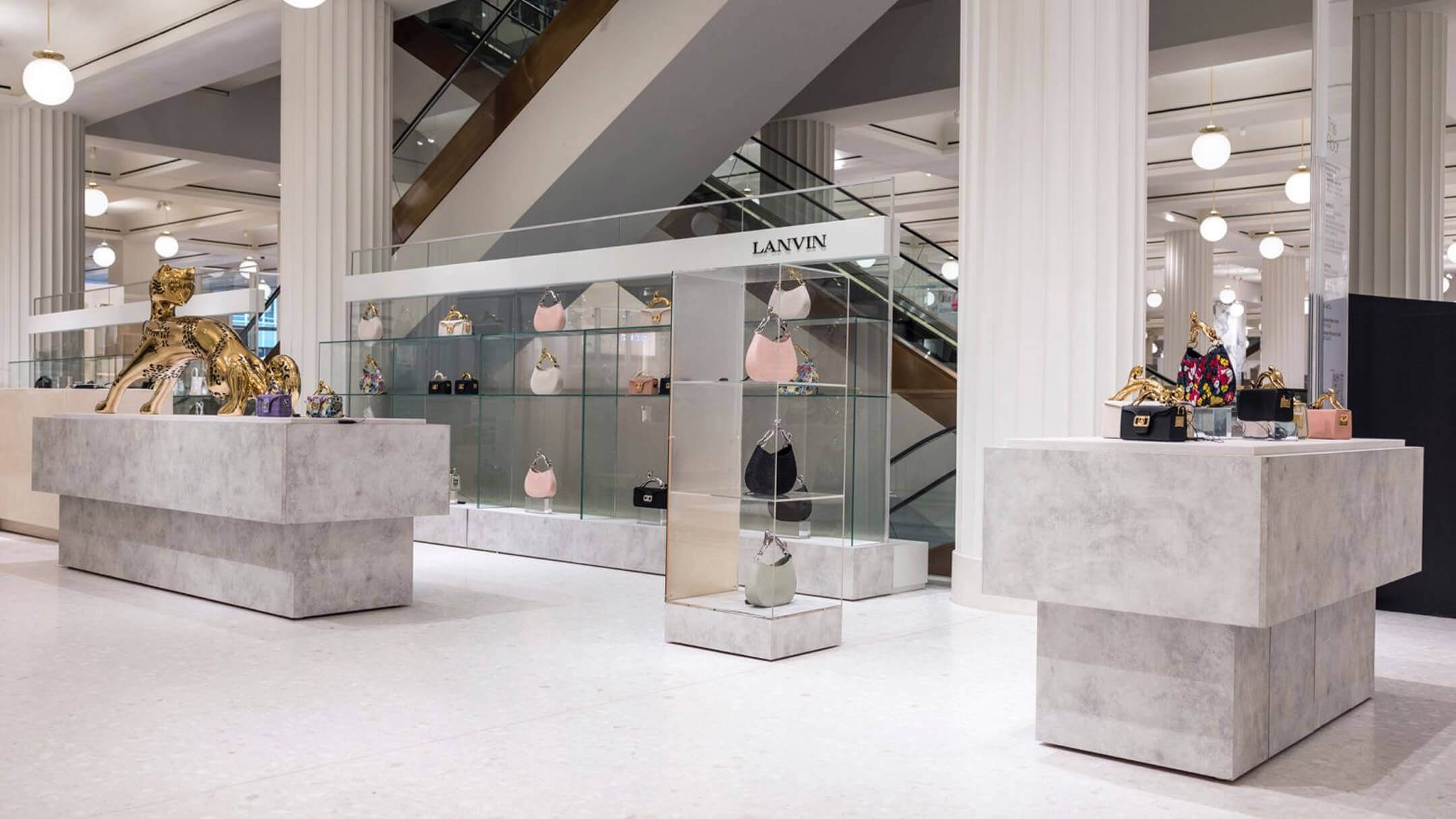 Lanvin retail display in a high-end department store, showcasing designer handbags on minimalist stone tables and glass shelving, with a gold sculpture centerpiece.